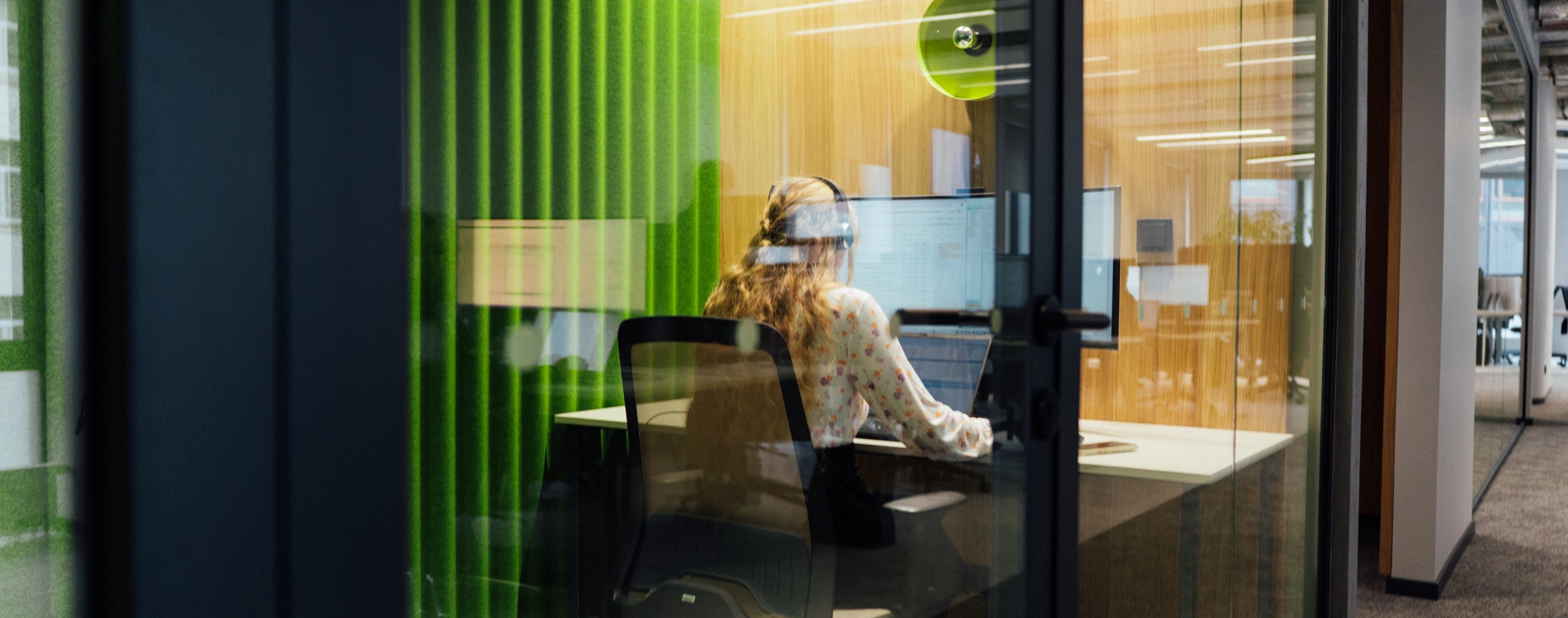 Person wearing a headset works at a desk inside a glass-walled office booth, facing computer monitors. The workspace features green acoustic wall panels, wood finishes, and modern office lighting.