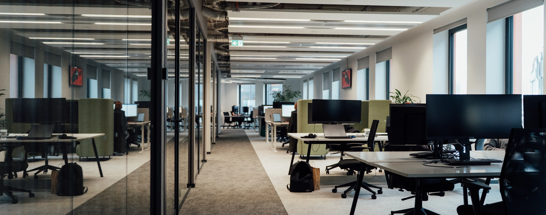 Modern open-plan office space with workstations, computer monitors, meeting rooms, and large windows providing natural light.