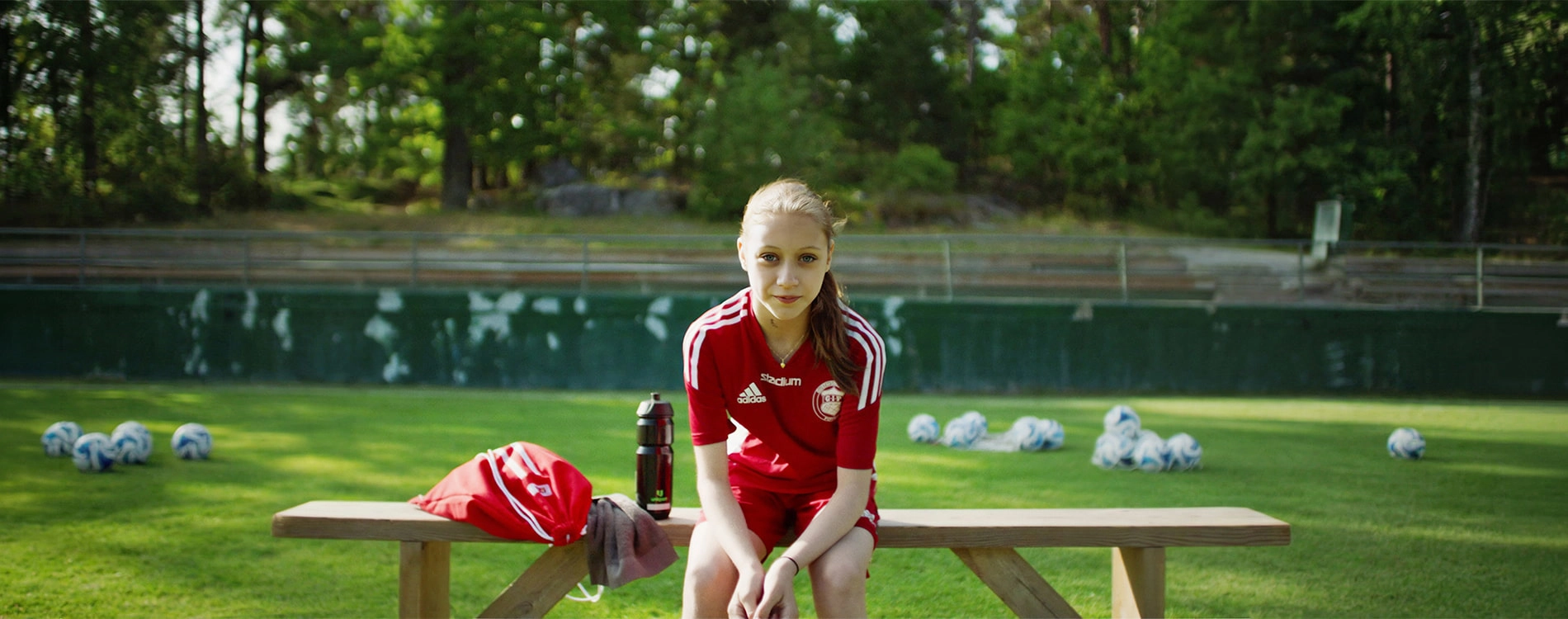 Young football player in a red sports uniform sitting on a football field during training, with footballs and a water bottle nearby.